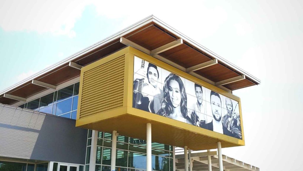 A modern, geometric and plate-glass museum rises in front of a clear blue sky. The front of the building is gray and yellow and covered with super imposed images of celebrity singers.