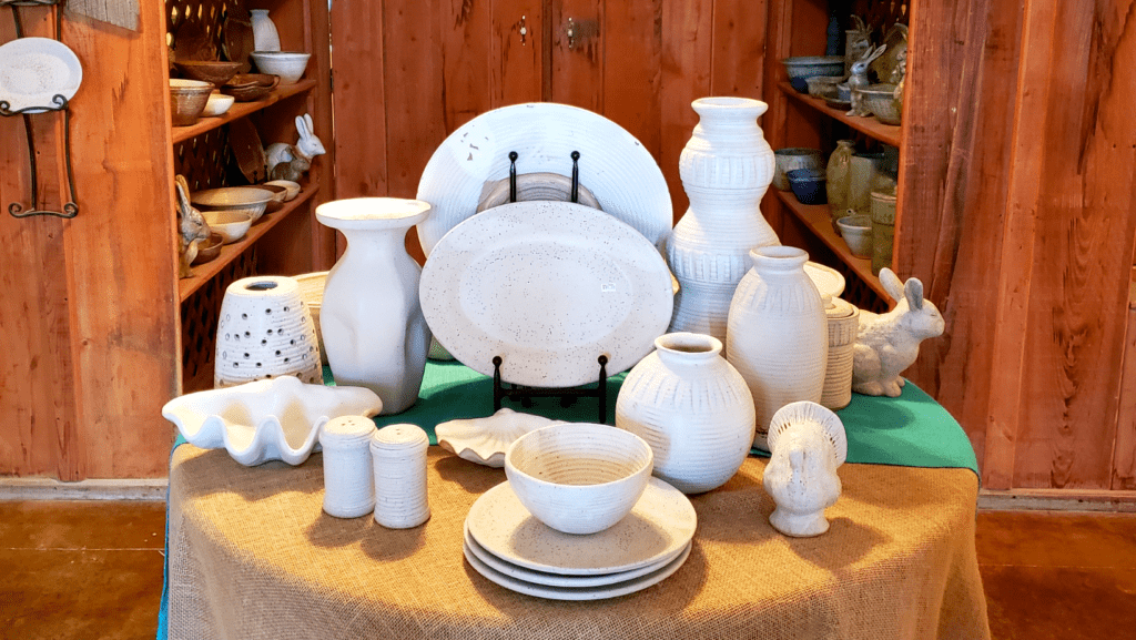 Cotton white platters, bowls, plates, and vases sit on a table covered by a burlap tablecloth.