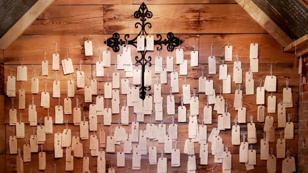 A wooden wall with a wrought iron cross covered with paper tags bearing handwritten prayers.